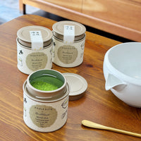Three round metal canisters of Konikafir matcha tea with beige lids and labels are arranged on a wooden table, with one canister open to show bright green matcha powder inside. A white ceramic bowl and a gold spoon rest to the right, with a wooden cabinet in the background.