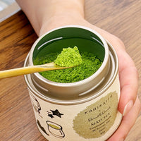 A hand holds an open round tin of Kohiraito Rooibos Matcha tea powder, showing the bright green powder and a wooden spoon, against a warm wooden tabletop background.